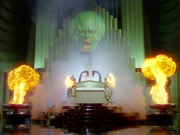 A still from The Wizard of Oz showing the image of the wizard projected green and large with flames all around.